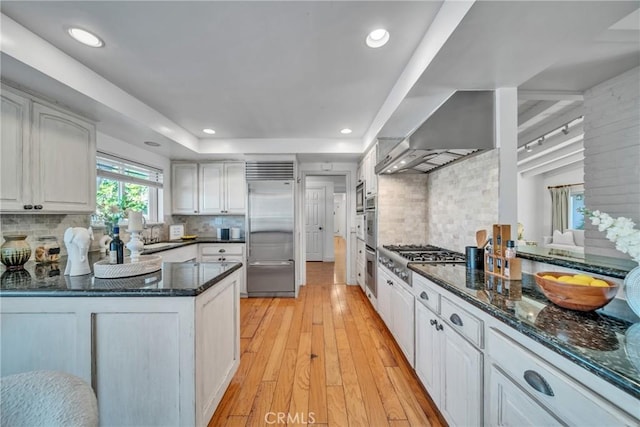 kitchen with white cabinets, wall chimney range hood, stainless steel appliances, backsplash, and light hardwood / wood-style flooring