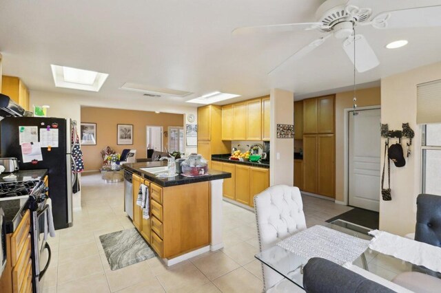 kitchen featuring ceiling fan, appliances with stainless steel finishes, an island with sink, light tile patterned floors, and light brown cabinetry