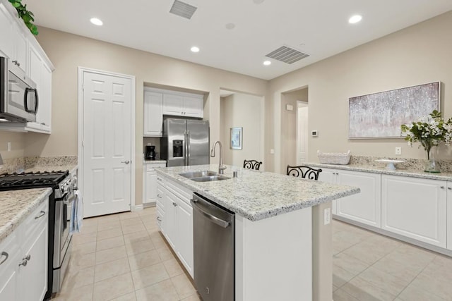 kitchen with an island with sink, stainless steel appliances, white cabinets, light stone counters, and sink