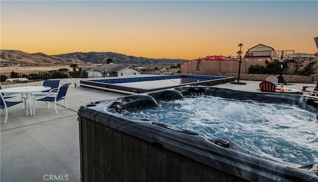 pool at dusk with a mountain view, a patio, outdoor dining area, and a hot tub