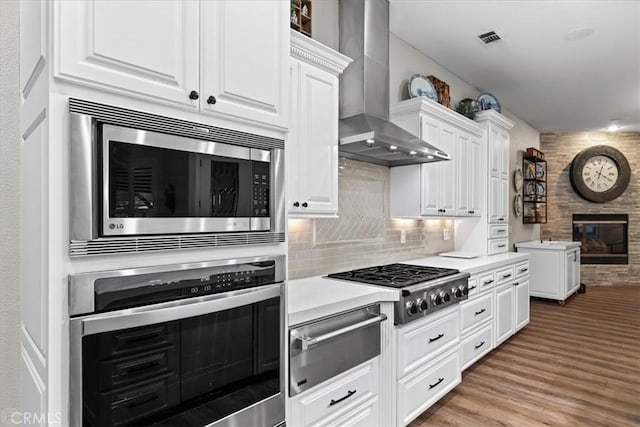 kitchen featuring white cabinetry, appliances with stainless steel finishes, tasteful backsplash, a fireplace, and wall chimney range hood