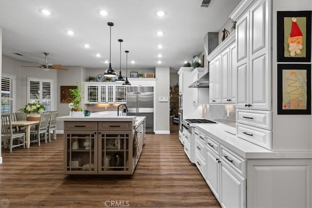 kitchen with ceiling fan, wall chimney range hood, pendant lighting, an island with sink, and white cabinets