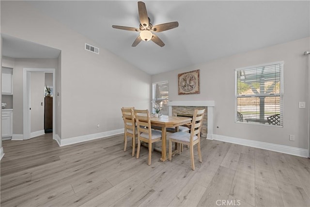 dining space with ceiling fan, light hardwood / wood-style flooring, a stone fireplace, and vaulted ceiling