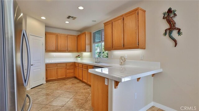 kitchen with tile counters, sink, stainless steel refrigerator, kitchen peninsula, and light tile patterned flooring