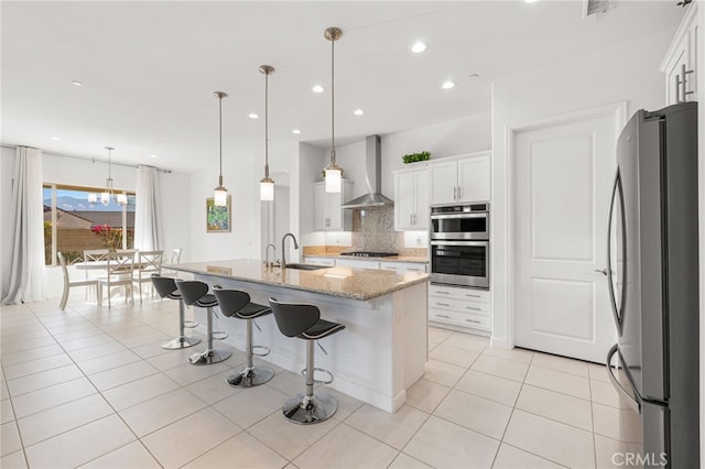 kitchen featuring appliances with stainless steel finishes, hanging light fixtures, wall chimney exhaust hood, white cabinetry, and a kitchen island with sink