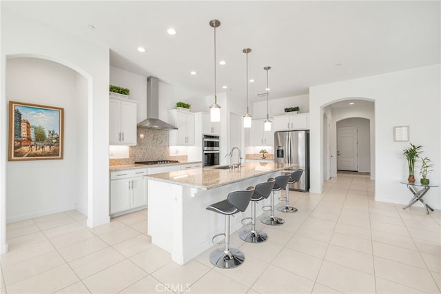 kitchen with a center island with sink, hanging light fixtures, stainless steel appliances, wall chimney range hood, and white cabinetry