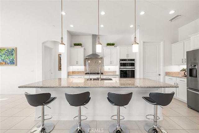 kitchen with hanging light fixtures, stainless steel appliances, white cabinets, wall chimney range hood, and a spacious island