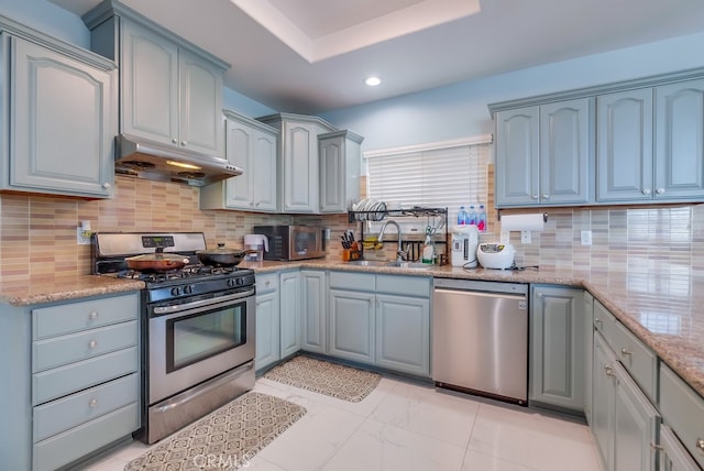 kitchen with light stone counters, under cabinet range hood, stainless steel appliances, a sink, and tasteful backsplash