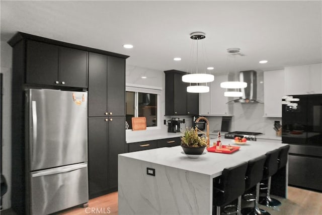 kitchen featuring wall chimney exhaust hood, decorative light fixtures, black refrigerator, an island with sink, and stainless steel refrigerator