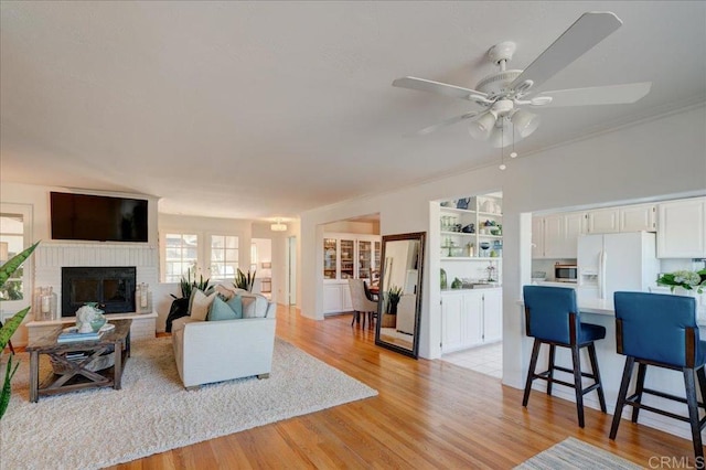 living room with built in features, ceiling fan, ornamental molding, a brick fireplace, and light wood-type flooring