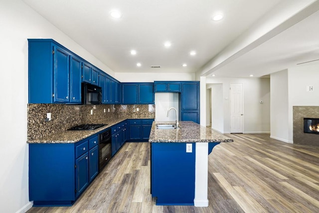 kitchen with black appliances, dark stone counters, backsplash, a center island with sink, and blue cabinets