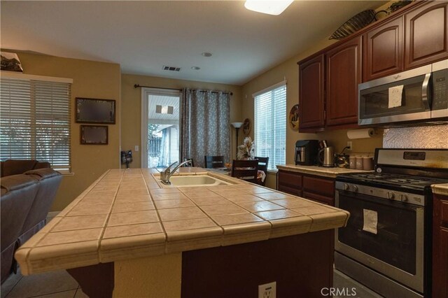 kitchen with tile patterned floors, tile countertops, a center island with sink, sink, and stainless steel appliances