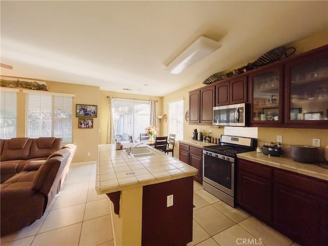 kitchen featuring light tile patterned floors, tile counters, glass insert cabinets, open floor plan, and stainless steel appliances