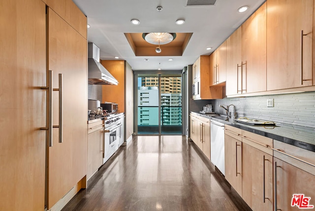 kitchen featuring light brown cabinets, appliances with stainless steel finishes, wall chimney range hood, and a tray ceiling