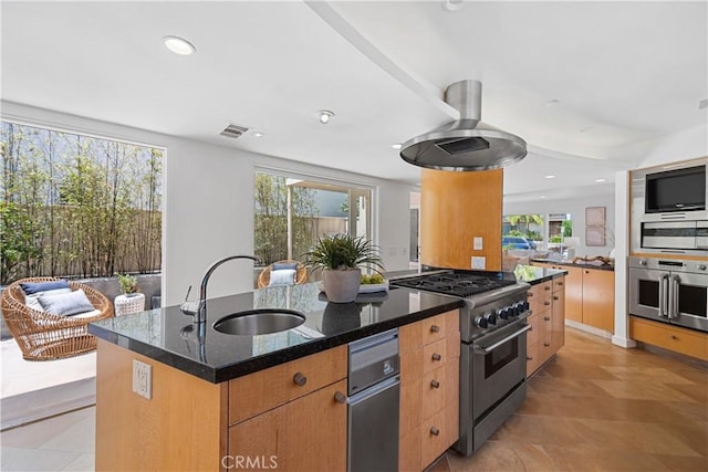 kitchen featuring stainless steel appliances, a healthy amount of sunlight, a kitchen island with sink, dark stone countertops, and sink