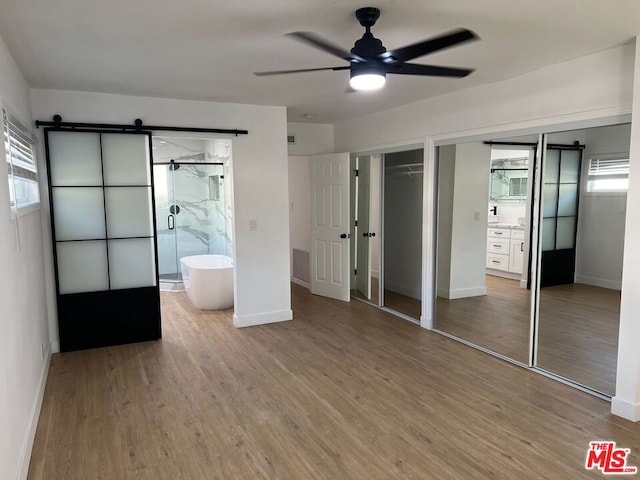 unfurnished bedroom featuring ceiling fan, connected bathroom, wood-type flooring, and a barn door