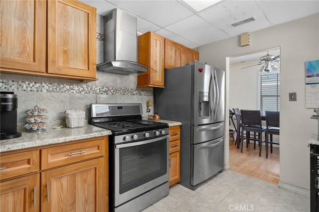 kitchen with ceiling fan, backsplash, wall chimney range hood, appliances with stainless steel finishes, and light stone countertops