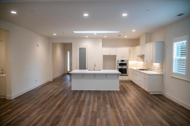 kitchen with white cabinetry, dark hardwood / wood-style flooring, double oven, a kitchen island with sink, and gas stovetop