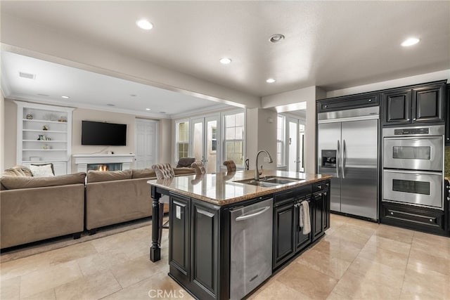 kitchen featuring appliances with stainless steel finishes, built in shelves, sink, a kitchen island with sink, and light stone counters