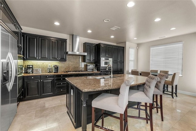 kitchen with an island with sink, a breakfast bar area, appliances with stainless steel finishes, wall chimney range hood, and sink