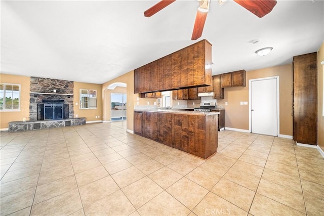 kitchen featuring light tile patterned floors, kitchen peninsula, a stone fireplace, ceiling fan, and stainless steel range oven