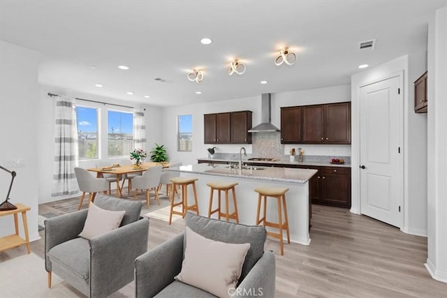 kitchen featuring wall chimney range hood, light hardwood / wood-style floors, a kitchen bar, a kitchen island with sink, and dark brown cabinets