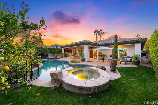 pool at dusk with a pool with connected hot tub, a patio, fence, and a lawn