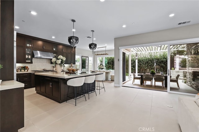 kitchen featuring range hood, decorative backsplash, hanging light fixtures, an island with sink, and dark brown cabinets