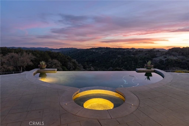 pool at dusk featuring a patio and a view of trees