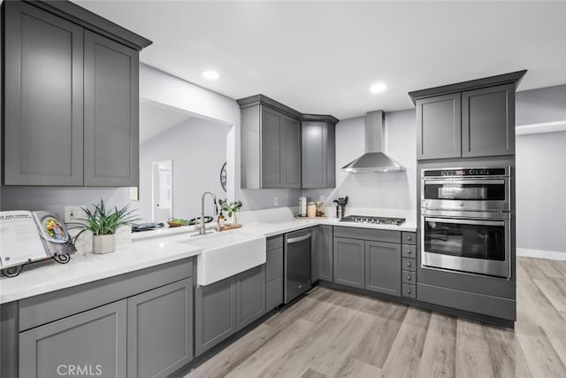 kitchen featuring appliances with stainless steel finishes, sink, gray cabinetry, wall chimney range hood, and light hardwood / wood-style flooring