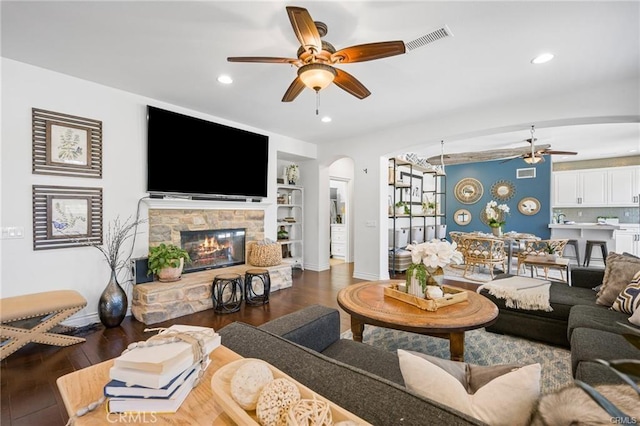 living room featuring built in shelves, ceiling fan, a stone fireplace, and dark hardwood / wood-style floors