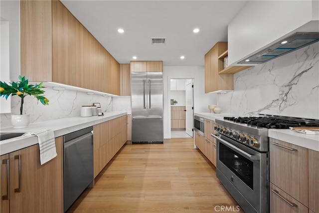 kitchen featuring wall chimney exhaust hood, tasteful backsplash, built in appliances, light brown cabinets, and light wood-type flooring