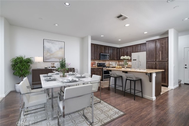 kitchen featuring a kitchen bar, a center island with sink, appliances with stainless steel finishes, decorative backsplash, and dark wood-type flooring