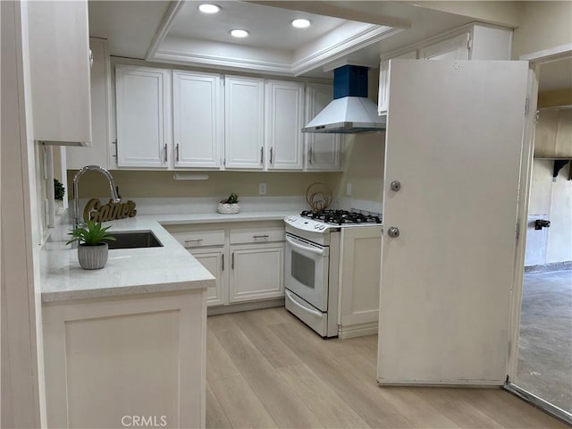 kitchen with a tray ceiling, white cabinets, white range with gas cooktop, wall chimney range hood, and a sink