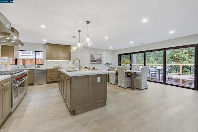 kitchen featuring appliances with stainless steel finishes, tasteful backsplash, an island with sink, hanging light fixtures, and wall chimney range hood