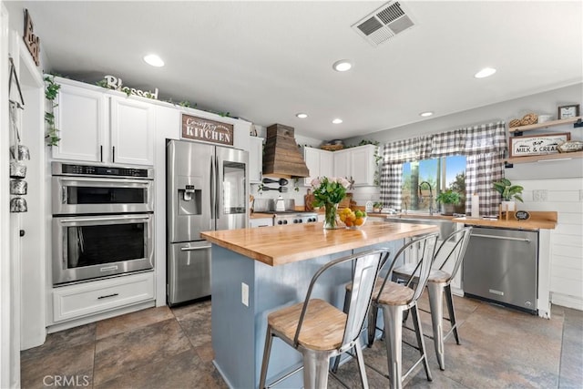 kitchen with a kitchen bar, white cabinetry, stainless steel appliances, butcher block countertops, and custom range hood