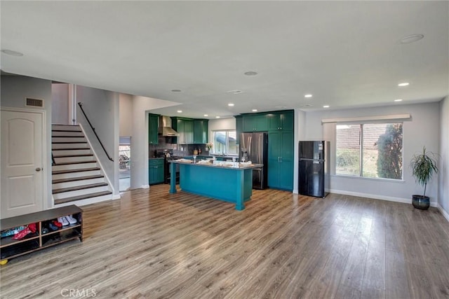 kitchen featuring wall chimney range hood, tasteful backsplash, appliances with stainless steel finishes, green cabinets, and light wood finished floors