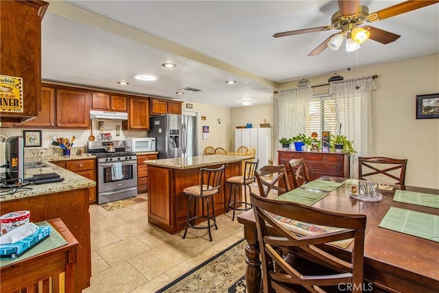 kitchen featuring light stone countertops, appliances with stainless steel finishes, a kitchen island, a kitchen bar, and ceiling fan