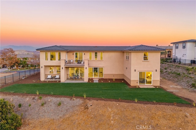 back house at dusk with a balcony, a patio area, and a lawn