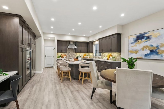 kitchen with light hardwood / wood-style floors, a kitchen bar, wall chimney range hood, dark brown cabinetry, and a center island