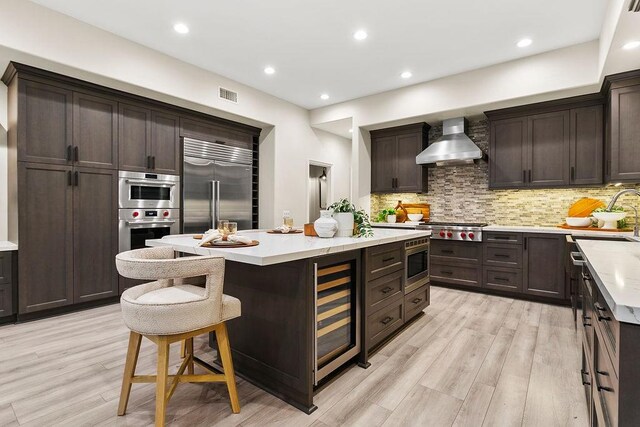 kitchen with a center island, wall chimney exhaust hood, built in appliances, wine cooler, and decorative backsplash