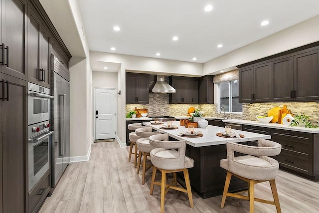 kitchen with light hardwood / wood-style floors, a kitchen island, a breakfast bar area, stainless steel appliances, and wall chimney exhaust hood