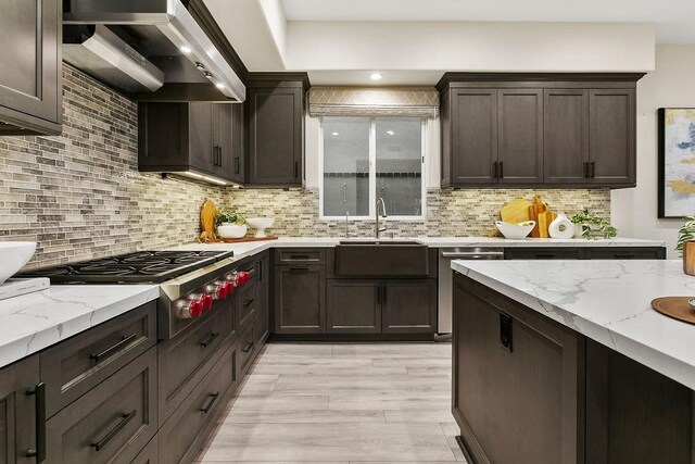 kitchen with appliances with stainless steel finishes, wall chimney range hood, decorative backsplash, sink, and light hardwood / wood-style flooring