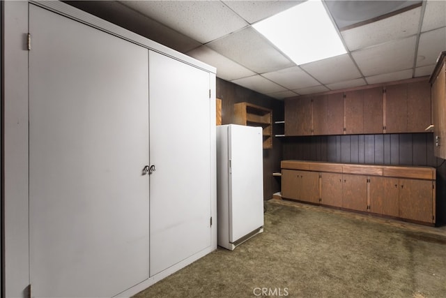 basement featuring a paneled ceiling, wooden walls, white refrigerator, and dark carpet