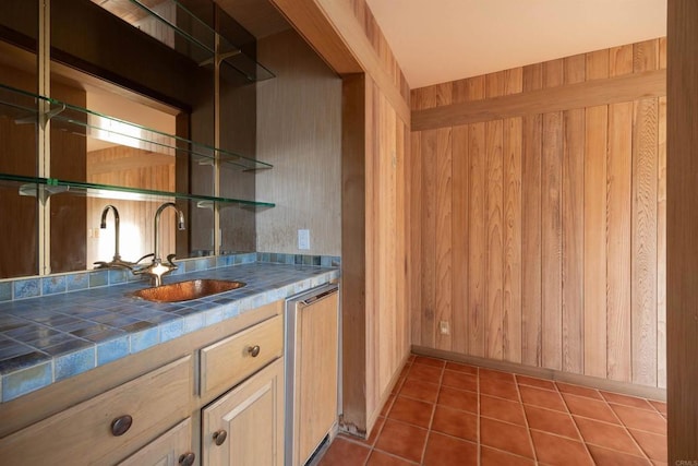 bar with sink, tile counters, wooden walls, and light brown cabinets