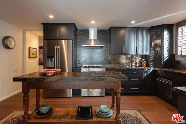 kitchen with wood-type flooring, wall chimney range hood, stainless steel appliances, dark stone countertops, and tasteful backsplash