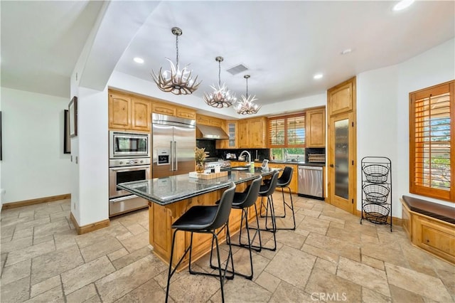 kitchen with wall chimney exhaust hood, a kitchen island, built in appliances, dark stone countertops, and a notable chandelier