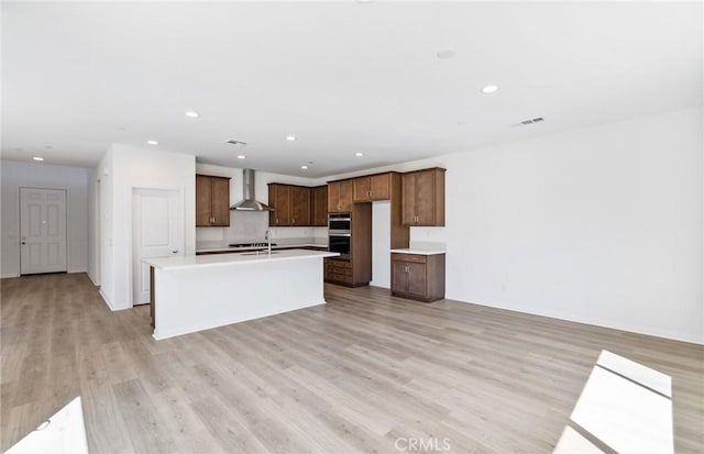 kitchen featuring stainless steel double oven, light hardwood / wood-style floors, a kitchen island with sink, and wall chimney range hood