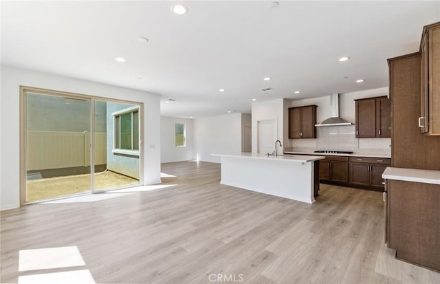 kitchen featuring an island with sink, sink, black gas stovetop, wall chimney range hood, and light hardwood / wood-style flooring