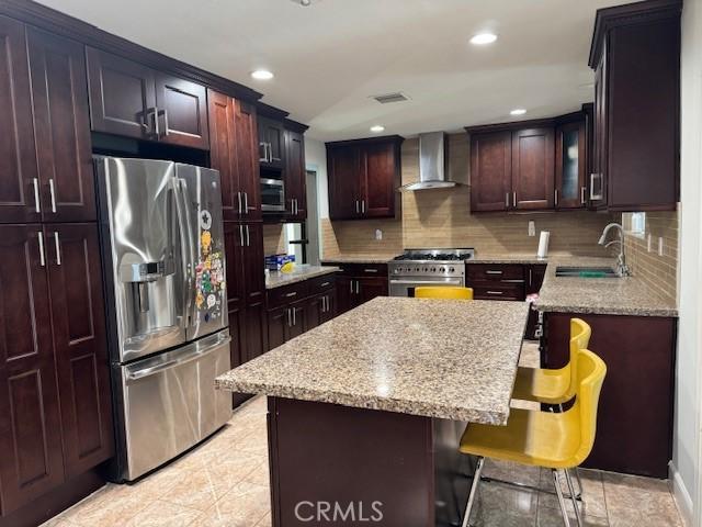 kitchen featuring light stone counters, appliances with stainless steel finishes, a sink, wall chimney range hood, and a kitchen breakfast bar